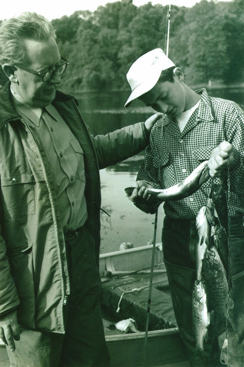 A historical black and white photo of two men standing near a boat on the shoreline of a lake. A young man inspects one fish from a string of fish he just successfully caught on the lake. The other, older man pats his shoulder and looks at the fish too. In the background are trees along the shoreline.