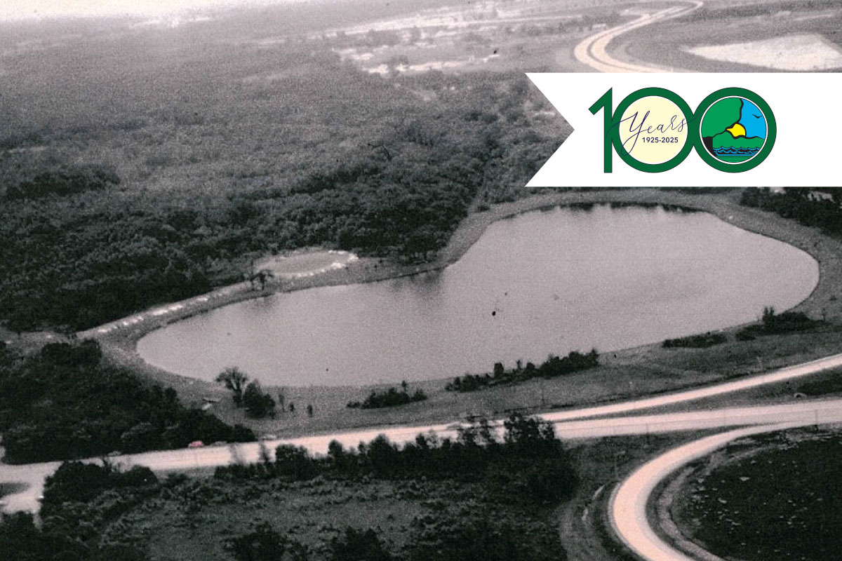 A historical black and white aerial view of a barrow pit lake on Cook County Forest Preserve property. Overlaying the photo on the top right is the logo for the Illinois Department of Natural Resources 100th anniversary.