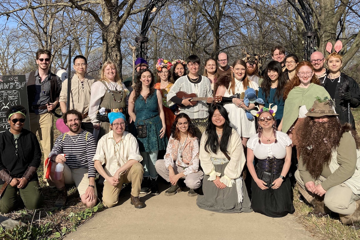 A group of people pose together. Everyone is wearing fantasy style costumes including some knights and elves. In the background are trees.