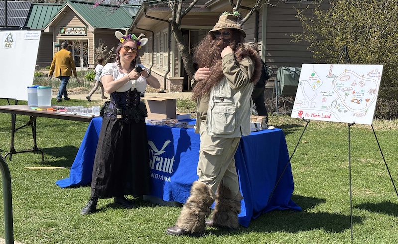 On a mowed lawn two people in medieval and fantasy costumes stand at a table with posters. In the background is a building with a sign with words saying, "Anita Purves Nature Center."