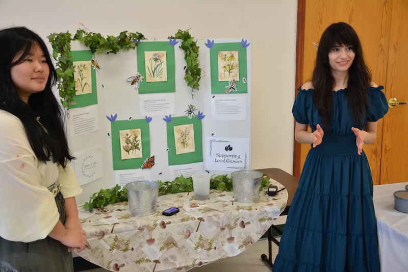 Two women in medieval elven costumes stand on either of a table. On top of the table is a poster board about supporting native plants and ecosystems.
