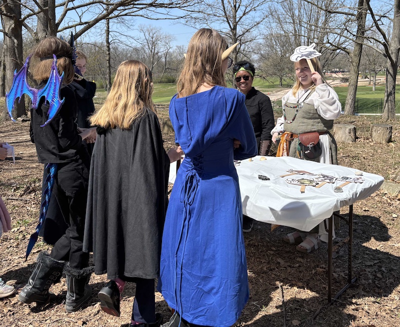 A group of people in medieval and fantasy costumes stand around a table. In the background are trees and a mowed lawn.
