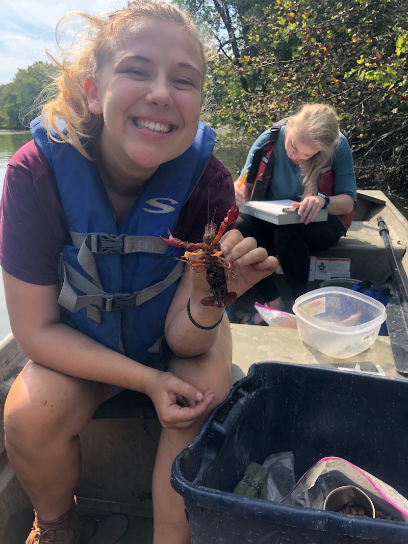 Two students sit in a boat along the shoreline of a river. One of the students holds a crayfish while the other student records data. In the background are trees filling the river shoreline.