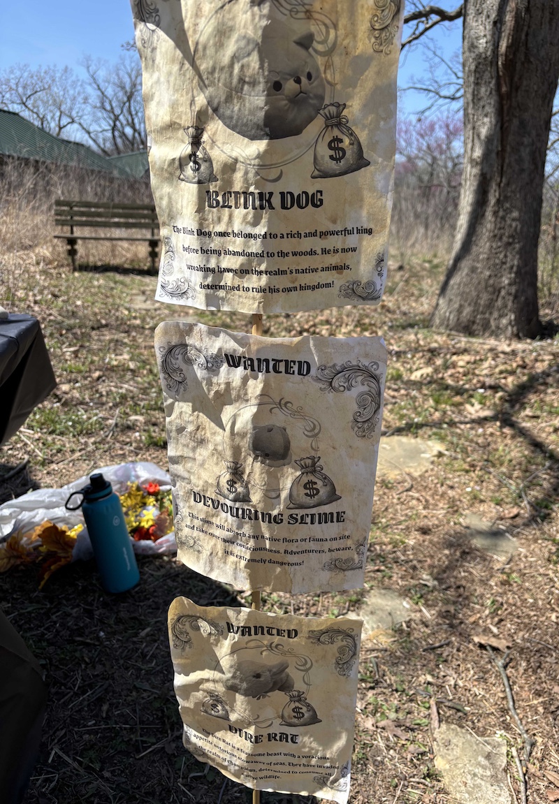 Three signs in aged brown paper feature photos of fantasy animals with the word "Wanted" at the top and a description of the fantasy animals underneath the images. In the background is a grassy area near a building with trees. A bright blue sky is overhead.