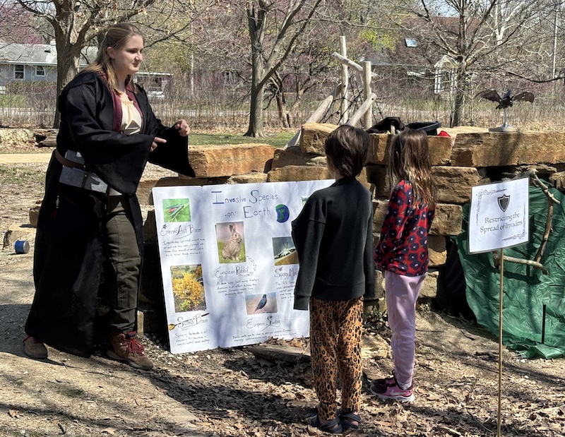 A woman in a medieval wizard costume talks to two young children. In the background a poster is resting on a stone bridge.