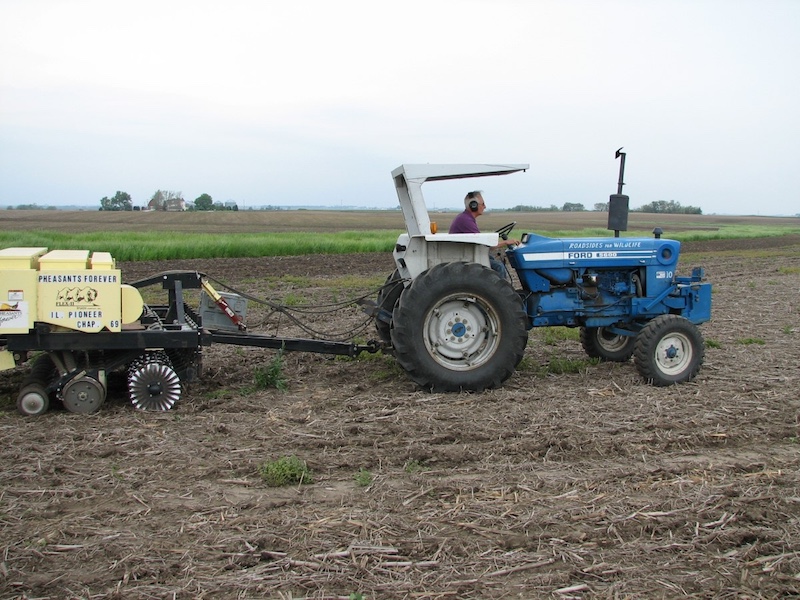 A man plants seeds in an agricultural field as he drives a blue tractor with a planter attached. 