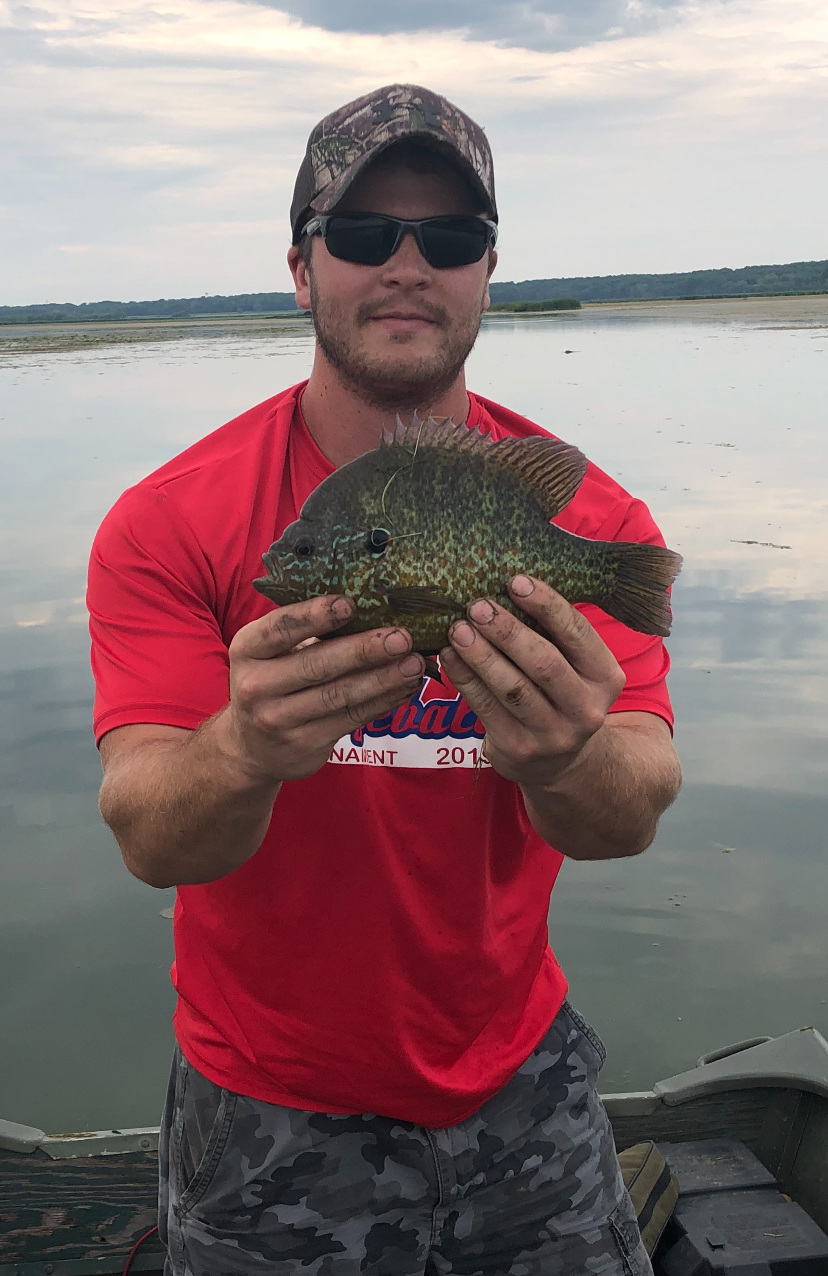 A man wearing a red shirt and a camouflage ball cap stands in a boat on a freshwater lake and holds up in front of him a green fish with both hands.