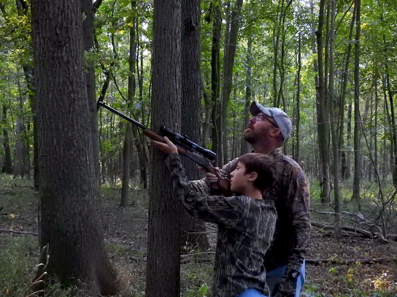 A boy in camouflage gear aims a small rifle while resting the barrel on a tree trunk towards a squirrel in a woodland canopy. An adult hunter stands close and mentors the young hunter through the shot. The adult hunter wears camouflage gear and a ball cap. In the background are trees and understory plants.