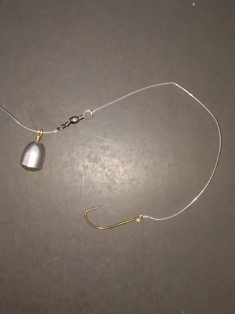 On top of a gray surface rests a fishing hook on a microfilament line with a fishing weight attached to one end.