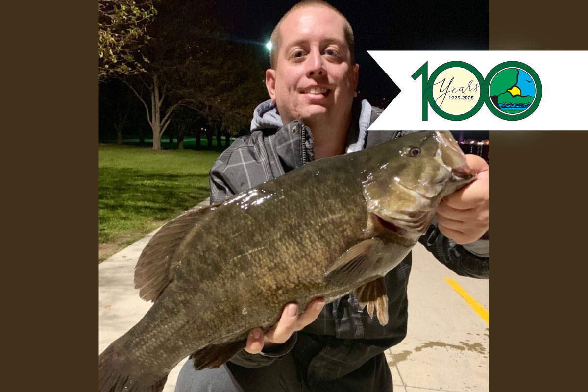 At night while kneeling on a concrete sidewalk, a male angler holds up with both hands a large green fish. Overlapping the man holding the fish on the top right is the Illinois Department of Natural Resources 100th Anniversary logo.