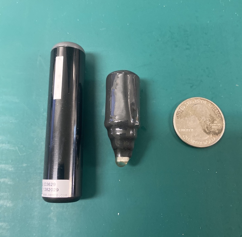 Two transmitters just slightly larger than a quarter rest on a green surface. One of the transmitters is shaped like a cylinder, and the other is shaped like a cylinder but with a point at one end. Both transmitters are black and dark gray.