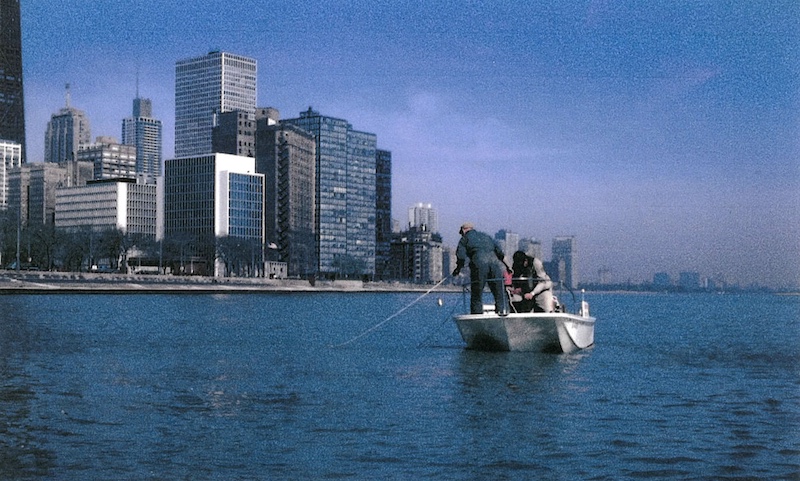 A historical photo of two biologists on a boat netting for fish in Lake Michigan off the Chicago shoreline. Skyscrapers are in the background.