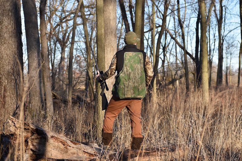 A hunter in camouflage gear pauses to listen in a fall woodland. The hunter holds a small rifle in his left hand. In the background blue sky filters through three trunks and limbs.