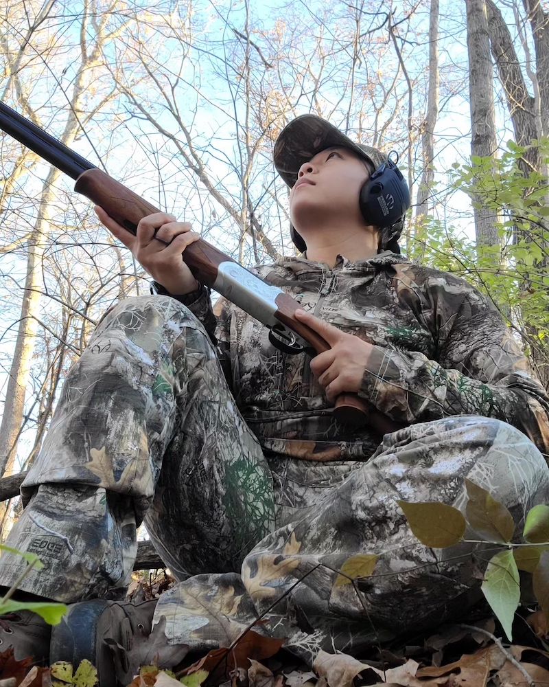 A hunter wearing camouflage sits on the ground in a woodland. She holds her shotgun at the ready to take quick aim.