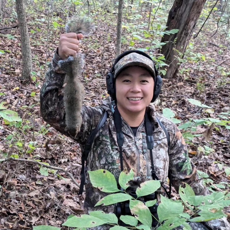 A hunter wearing camouflage gear stands proudly in a woodland, and holds up a successfully harvested gray squirrel.