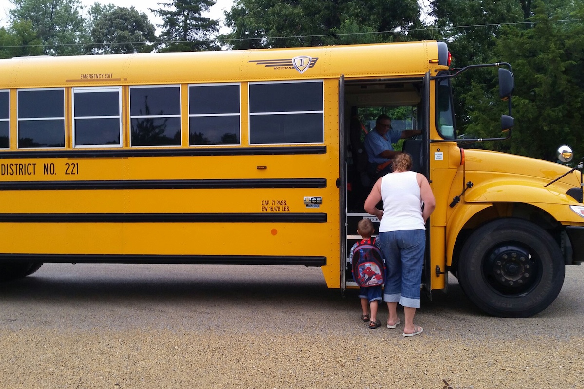 A mother helps her young boy up the stairs of a yellow school bus.