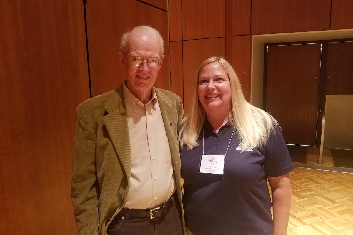 An elderly man in a tan corduroy blazer stands next to woman with long blonde hair and a navy blue polo shirt.