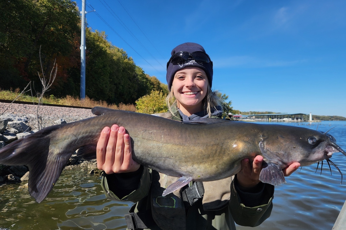 A young woman holds up with both hands a large gray, tan catfish while standing on a boat near the shoreline of a lake. In the background is a marina filled with boats.