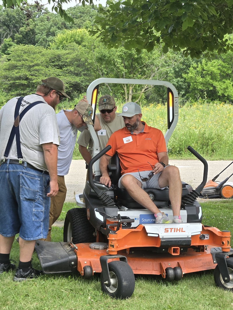 A man sits in an electric mower and shares information about the machine. Three other men look on and listen. In the background is a grassy field against a woodland.