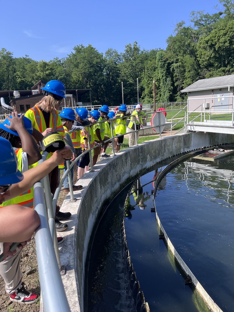 Students wearing blue hard hats and yellow high visibility vests observe the a clarification pool from behind a railing on top of a concrete retaining wall at wastewater treatment plant. In the background is a forest, and a bright blue sky is overhead.