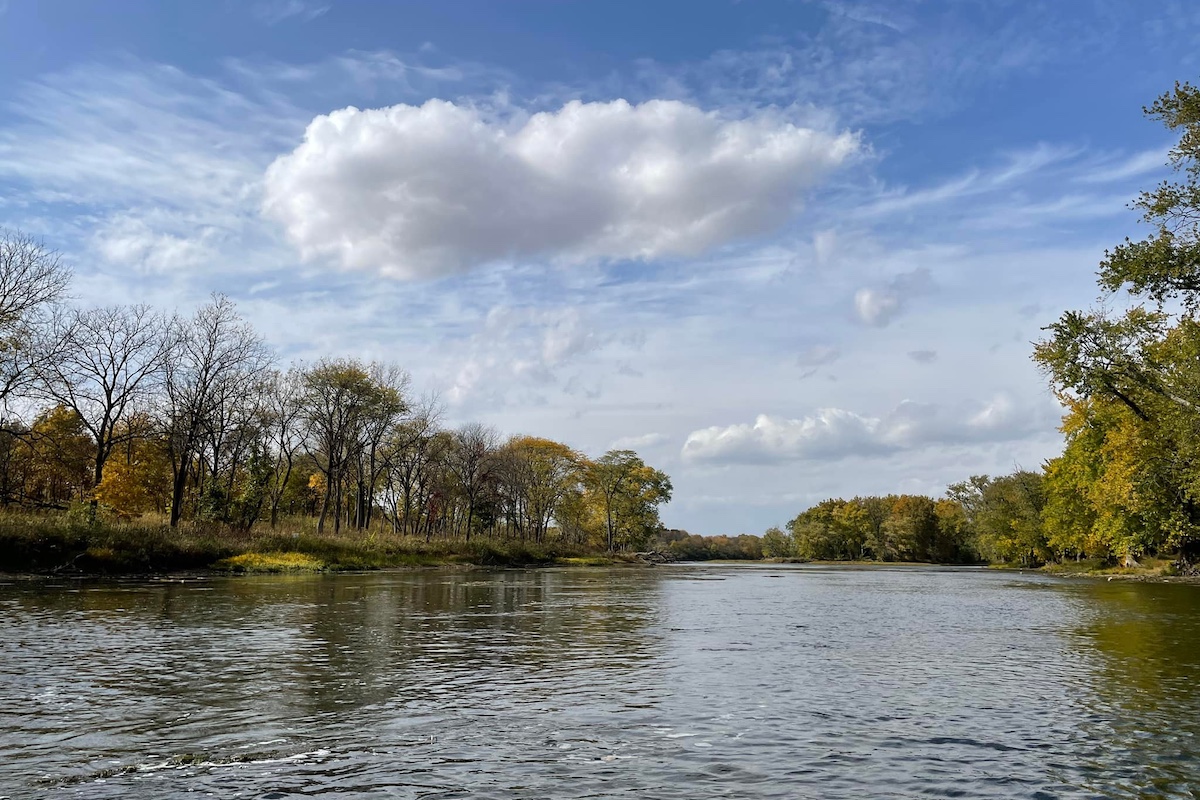 On a beautiful fall day, a partly cloudy sky is reflected in a river. On either side of the river is trees with fall foliage.