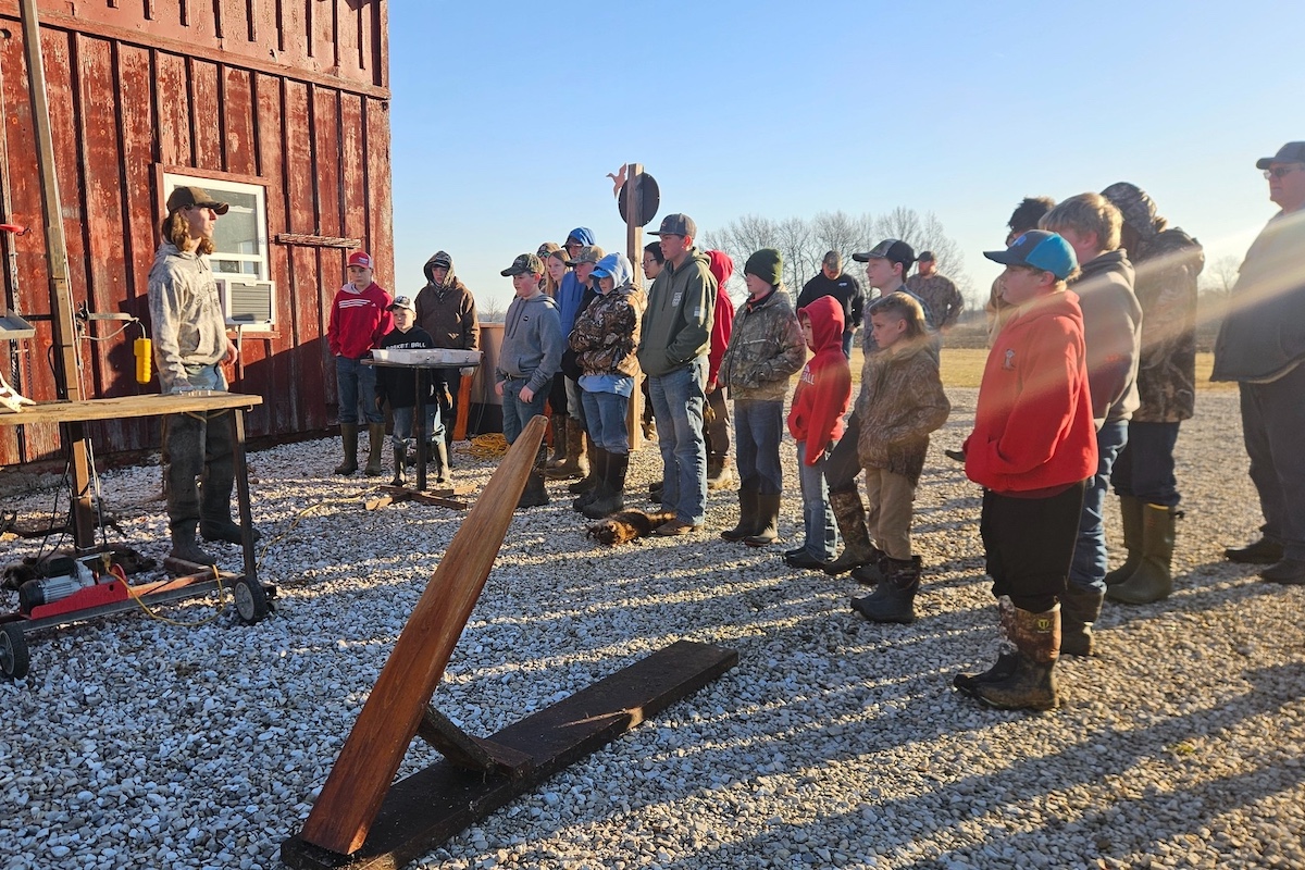 Under a bright blue sky, a group of people stand outside a red barn. A man presents to the group about fur handling while trapping. All are wearing warm sweatshirts or hoodies to stay warm in the chill fall air.