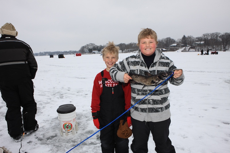 Two boys stand together on an ice covered lake. One of the boys holds up a successfully caught catfish. In the background are ice fisherman standing on the ice and attempting to catch a fish. Suburban houses can be seen on the hill that overlooks the shoreline of the lake.