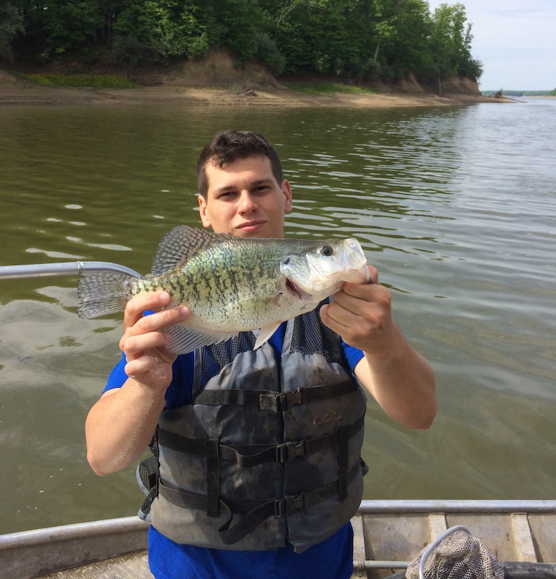 A man stands in a boat on a lake and holds up with both hands a medium size green fish with black spots. In the background is a shoreline filled with trees.