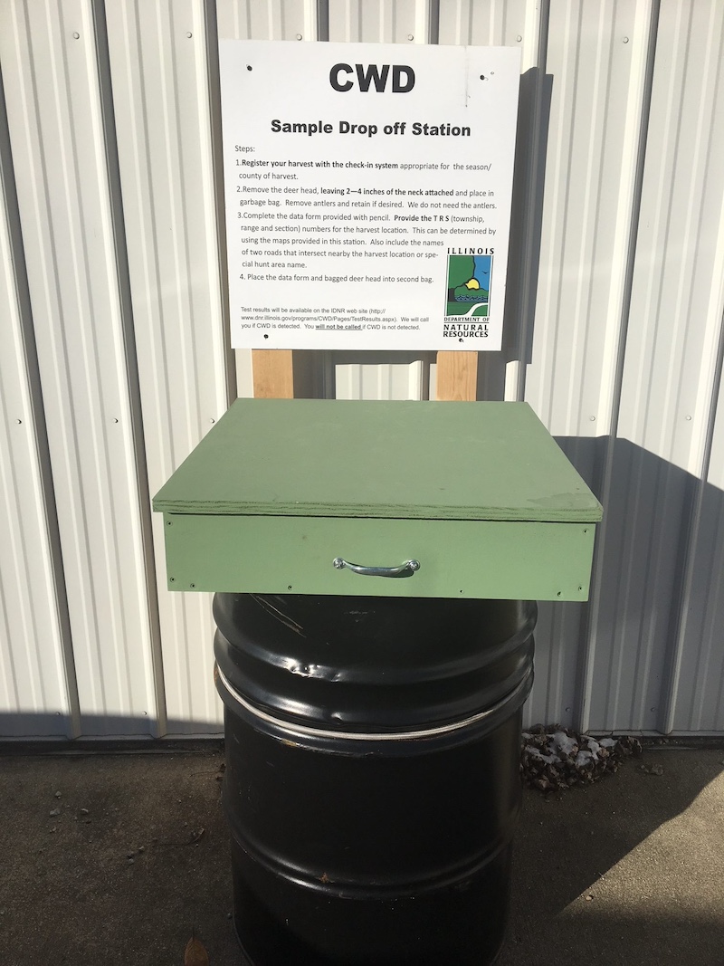 A green box installed on top of a black barrel positioned on the side of a metal sided building. A sign over the green box announces the box as a sample drop off station.
