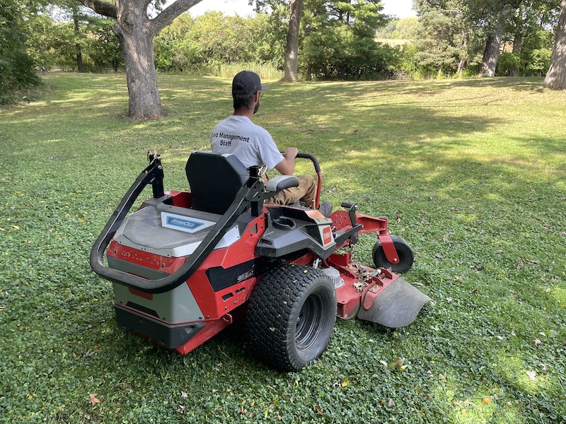 A technician at a state park mans an electric mower. In the background are trees providing shade on a sunny summer day.