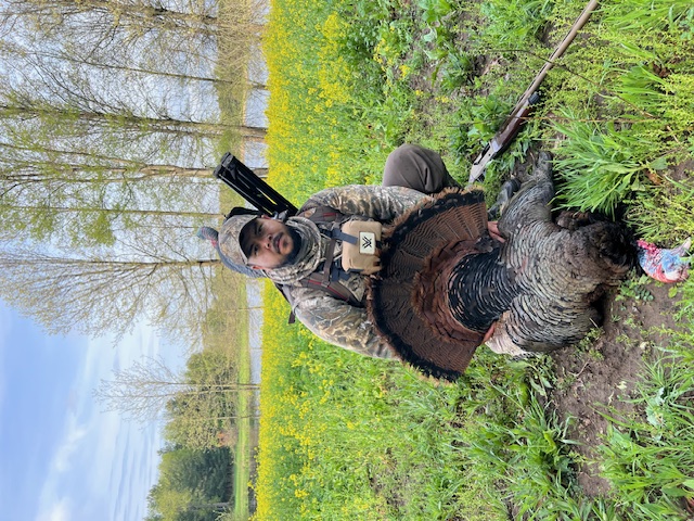 A hunter wearing camouflage gear in a spring grassland kneels on the ground and poses with a successfully harvested wild turkey. In the background is a pond surrounded by trees. A partly cloudy bright blue sky is overhead.