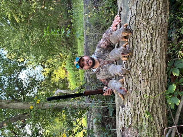 A hunter wearing camouflage gear poses behind a fallen log. Resting on the log in front of the hunter are four successfully harvested squirrels. In the background is a grassy area surrounded by trees.