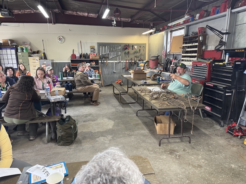 A man sits behind a table and talks about various types of traps to the group of women sitting at tables in front of the man. In the background is a workshop with different wrenches hanging on the wall, tool chests, and a work bench.