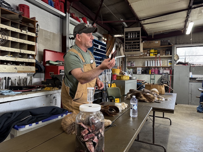 An instructor stands behind a table and talks about trapping equipment. In the background is a workshop with a work bench, tool chests, wall mounted cabinets, and shelving.