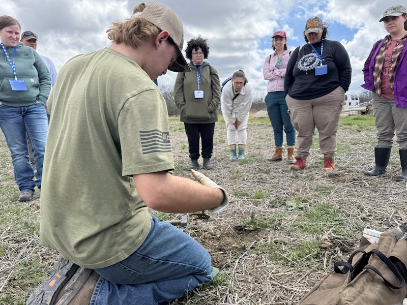 A group of women stand in a grassy field and watch an instructor give a trap setting demonstration. A partly cloudy sky is overhead.