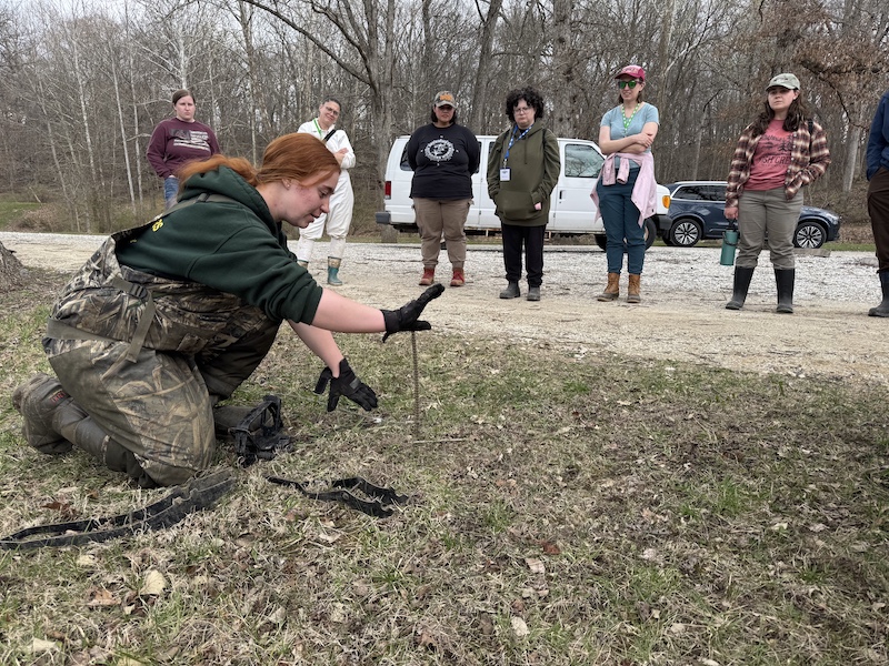 An instructor demonstrates how to set a trap to a group of women outdoors. In the background is a gravel parking lot in front of a forest.