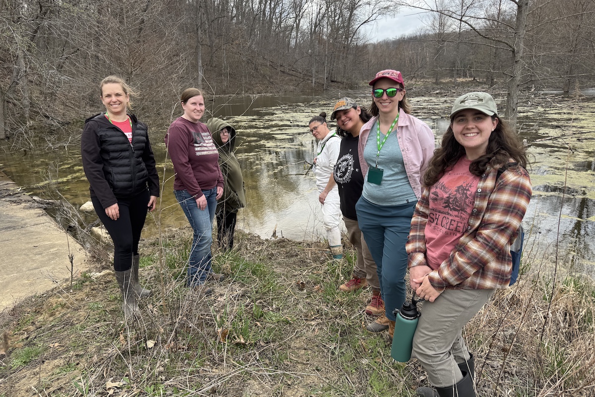 A group of women stand on the shoreline of a bottomland wetland in early spring. A hillside of trees surrounds the wetland on the left side. In the far background is a grassland and a woodland.