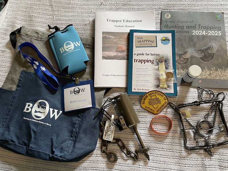 A photo of a collection of various trapping resources, a bag, drink koozie, two types of traps, a patch that reads, "Trapper Education Graduate," and a silicone bracelet. All the items are resting on top of a white knitted blanket.