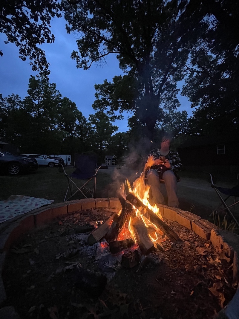 A campfire adds warmth to a chill autumn evening. A woman sits near the campfire and looks at her phone. Overhead are tree canopies silhouetted against a sky with fleeting evening light.