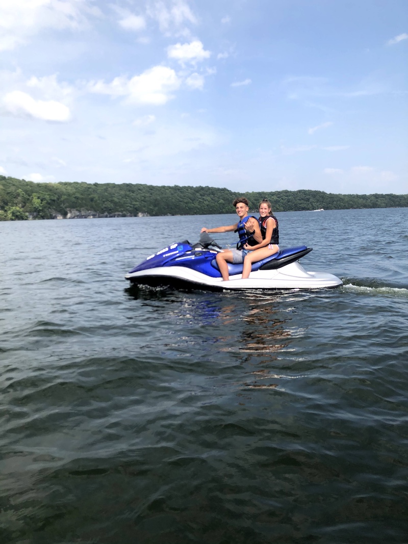 A teenage boy and girl ride a jet ski on a lake under a bright blue partly cloudy sky. In the background is a shoreline filled a dense forest, and a few rocky outcroppings emerge from the trees.