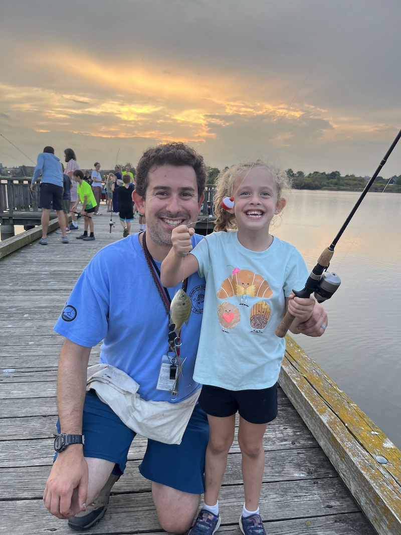 On a wooden dock over a lake, a man kneels next to a young girl. Both have beaming smiles while the girl holds up a fishing line with a small green fish caught on the hook. In her other hand, she holds up the fishing rod and reel.