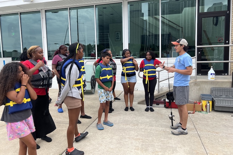 A group of young adults wear life-vests and stand in a group while listening to an instructor teach about fishing while holding a fishing rod and reel. In the background is building with large windows and a short staircase leading up to a door.