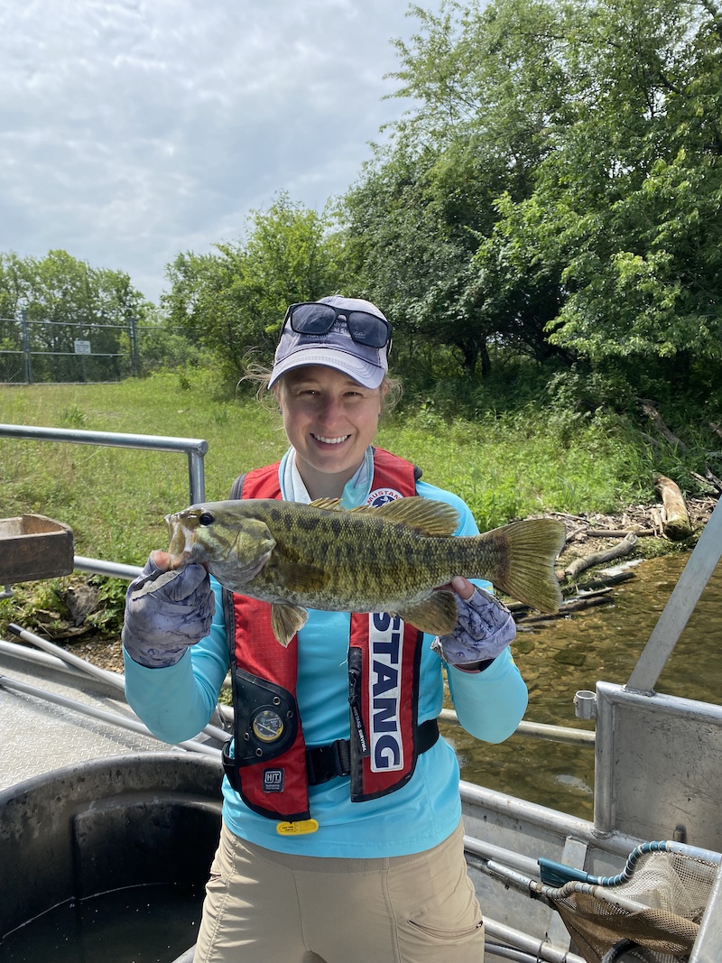 A woman stands on a boat and holds up with both hands a large green and black mottled fish. In the background is a grassy shoreline with bushes and trees to the right.