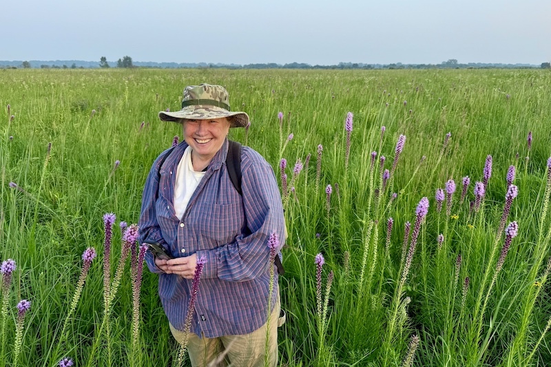 A woman stands in a green lush prairie. The woman is surrounded by purple prairie flowers.