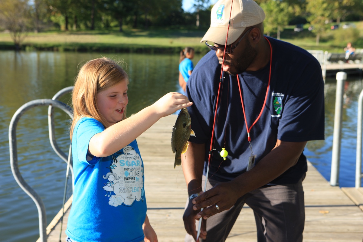 On a wooden dock over a freshwater pond, a biologist and a young girl who just caught a small green fish talk about taking the fish off the line. In the background are more people on the dock, and the green lawn beyond the shoreline.