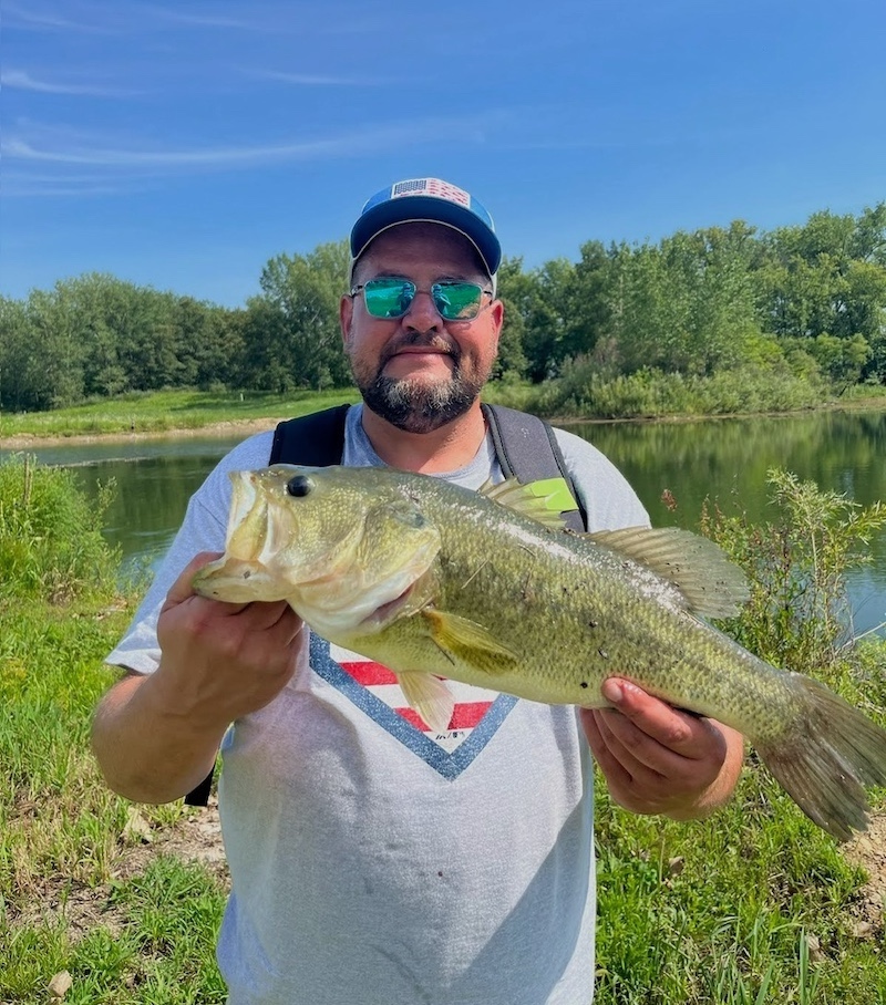A man holds up a large green fish with both hands. In the background is a lake reflecting a bright blue sky overhead.