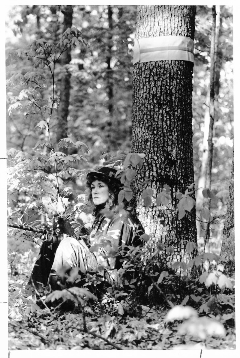A historical black and white photo of a hunter wearing camouflage gear sitting at the base of a tree with her back resting against the tree trunk in a woodland. She holds her camouflaged shotgun in her lap and is alert for signs of an approaching turkey.