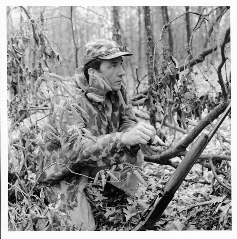 A historical black and white photo of a turkey hunter wearing camouflage gear kneeling in a woodland partially obscured by a fallen tree branch. The hunter is calling for wild turkeys with a slate turkey call. To his left, his shotgun is resting on the fallen tree branch.