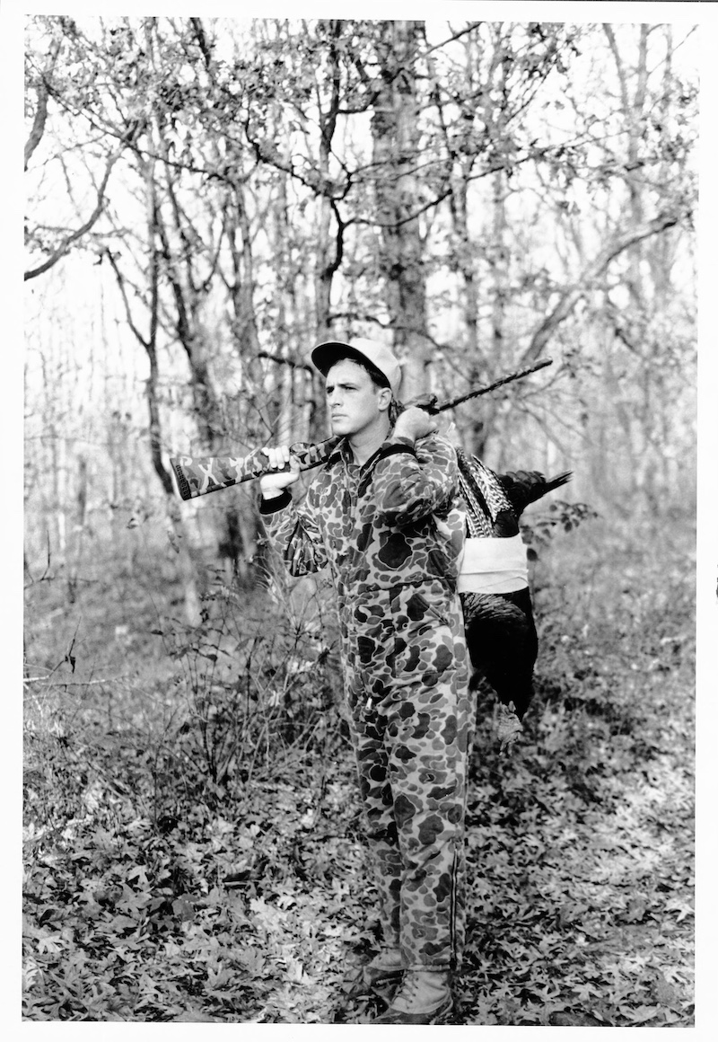 A historical black and white photo of a hunter wearing camouflage gear standing in a woodland with a successfully harvested wild turkey slung over his shoulder. In his right hand, he holds his shotgun and rests the barrel over his shoulder.