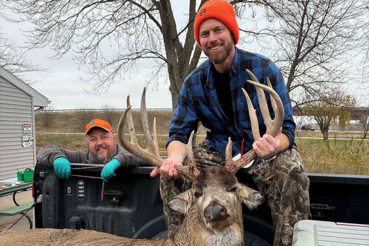 Two men pose next to a harvested adult male white-tailed deer in the bed of a pickup truck. One individual is sitting on the edge of bed of the pickup truck while he holds up the deer's large antlers. The man to the left leans with his arms over the side of the bed of the pick up truck. In the background is a highway overpass.